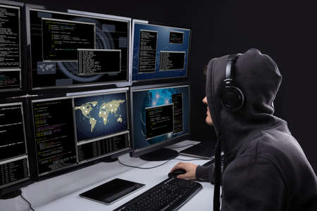 Rear View Of A Hacker Using Multiple Computers For Stealing Data In Office
