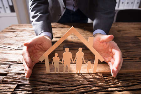 Businessperson's Hand Protecting House Model And Family Paper Cut Out On Wooden Deskの写真素材