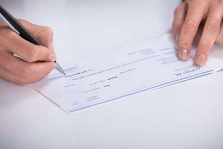 Close-up Of A Person's Hand Signing Cheque On Desk In Officeの写真素材