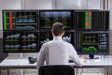 Side View Of A Young Male Stock Market Broker Analyzing Graphs On Multiple Computer Screens