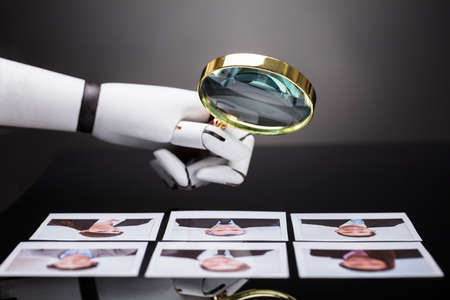 Close-up Of A Robot's Hand Looking At Candidate Photograph With Magnifying Glass