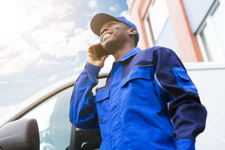 Low Angle View Of Happy Delivery Young Man Standing Near Van Talking On Cellphoneの写真素材