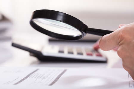 Elevated View Of Businessman Hand Examining Invoice With Magnifying Glass And Calculator In Officeの写真素材
