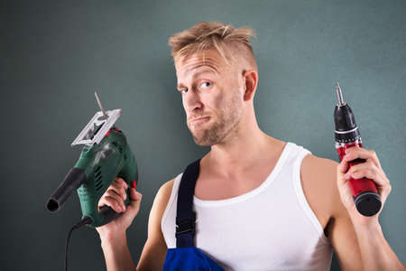 Portrait Of A Male Technician Holding Electric Drill And Screwdriver On Grey Background