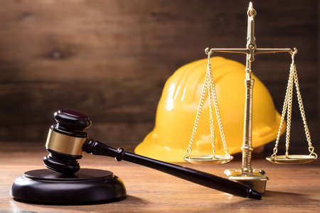 Judge Gavel In Front Of Yellow Safety Helmet And Golden Scale On The Wooden Table