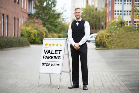 Portrait Of A Smiling Young Male Valet Standing Near Valet Parking Signの写真素材
