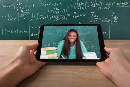 Close-up Of A Student's Hand Video Conferencing With Happy Female Teacher On Tablet Over Deskの写真素材