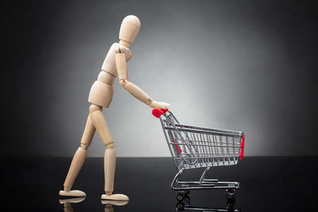 Side View Of A Wooden Dummy Pushing Shopping Cart On Grey Backgroundの写真素材