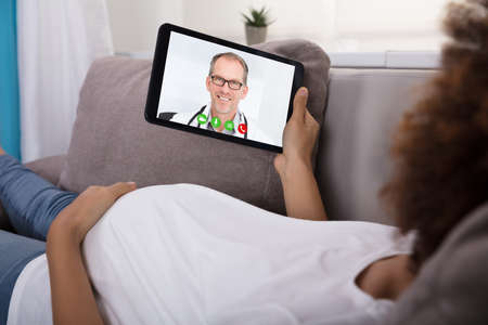Close-up Of Expecting Pregnant Woman Lying On Sofa Video Conferencing With Doctor On Digital Tablet