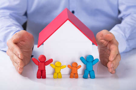 Close-up Of Man's Hand Protecting The House Model With Colorful Family On Deskの写真素材