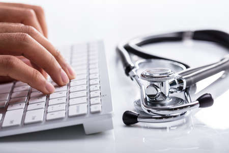 Doctor Typing On Computer Keyboard With Stethoscope On White Deskの写真素材