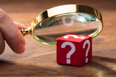 Close-up Of A Person Looking At Wooden Question Mark Red Block Through Magnifying Glass