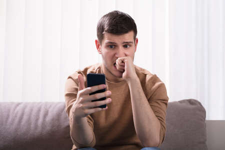 Worried Young Man Looking At Smartphone And Sitting In Front Of Sofa At Home