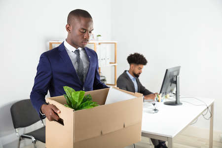 Businessman Moving Out With Cardboard Box From Office With Employee Working At Deskの写真素材