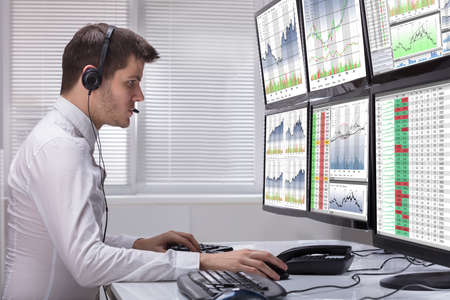 Side View Of A Young Male Stock Market Broker Analyzing Graphs On Multiple Computer Screens