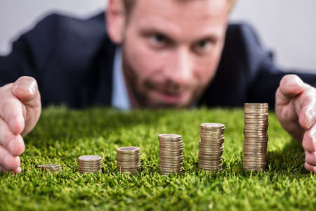 Close-up Of A Businessman's Hand Protecting Stacked Coins On Green Grassの写真素材