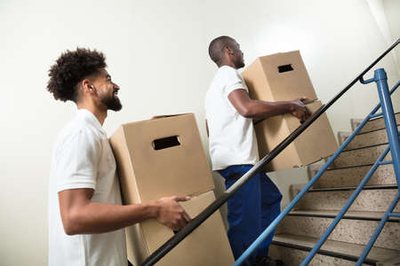 Two Young Male Worker Holding Cardboard Boxes While Climbing Steps At Homeの写真素材