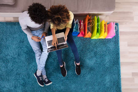 Young Couple Shopping Online On Laptop With Multi Colored Shopping Bags On Sofaの写真素材