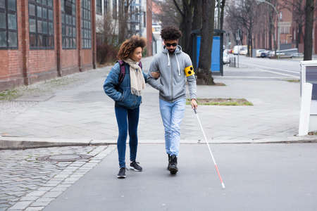 Young Woman Helping Blind Man With White Stick While Crossing Roadの写真素材