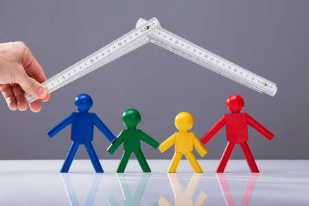 Businessperson's Hand Holding Measurement Tape Over Multi Colored Human Figures On White Deskの写真素材