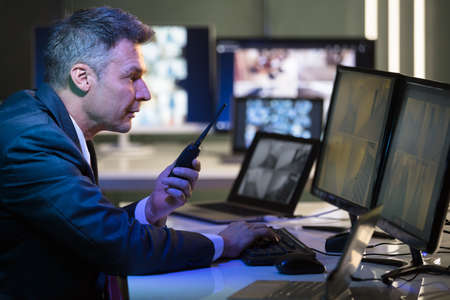 Businessman Talking On Walkie Talkie While Looking At CCTV Camera Footage On Multiple Computer...