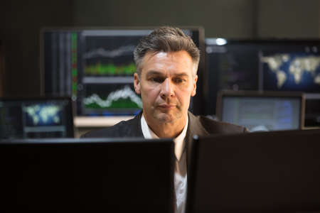 Mature Male Stock Market Broker Looking At Multiple Computer Screen In Office
