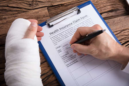 Close-up Of A Businessperson With Broken Arm Filling Health Insurance Claim Form On Wooden Desk