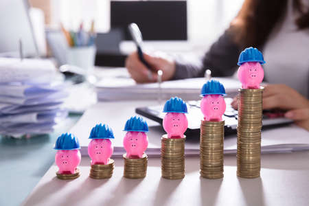 Close-up Of Pink Piggybank With Blue Hard Hat Over Increasing Stacked Coins At Workplace