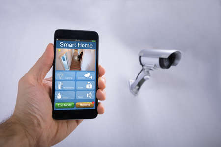 Human Hand Using Smart Home Application On Smartphone Near CCTV Camera Mounted On Wallの写真素材