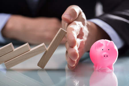 Close-up Of A Businessman's Hand Stopping Wooden Blocks From Falling On Piggybank Over Desk