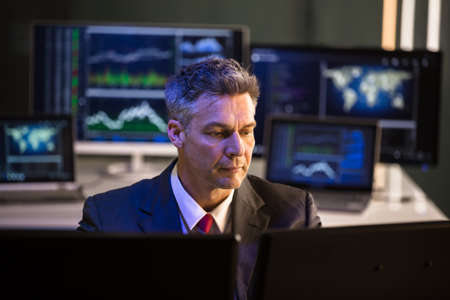 Mature Male Stock Market Broker Looking At Multiple Computer Screen In Office