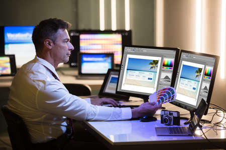Side View Of A Mature Male Designer Holding Colorful Swatch Working On Computer In Office