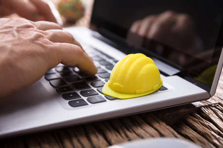 Close-up Of An Architecture's Hand Using Laptop With Yellow Hard Hat On Keypadの写真素材