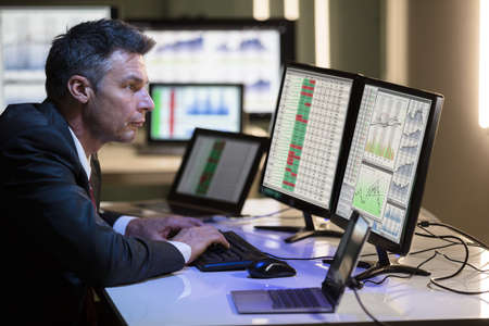 Side View Of A Mature Male Stock Market Broker Analyzing Graph On Multiple Computer Screen