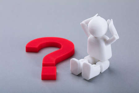Close-up Of A Human Figure Sitting Beside Red Question Mark Sign On Grey Background