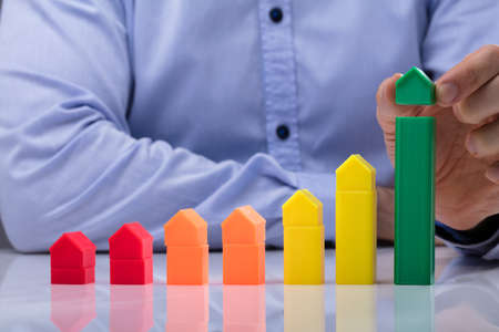 Person's Hand Placing Small Green House On Top Of Increasing Multi Colored Graph