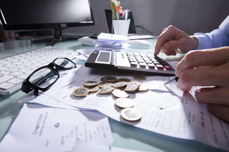 Businessperson's Hand Calculating Receipt With Coins And Eyeglasses On Desk