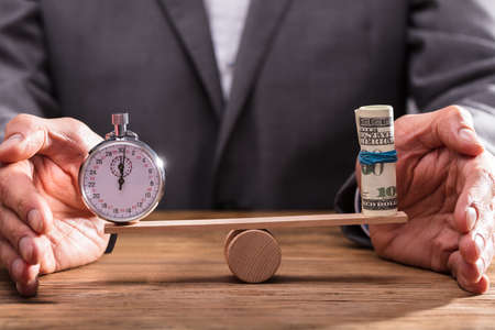 Close-up Of A Businessperson's Hand Protecting Balance Between Stopwatch And Rolled Up Banknotes On Seesawの写真素材