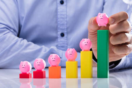 Close-up Of A Businessperson's Hand Placing Piggy Bank On Top Of Multi Colored Business Graphの写真素材