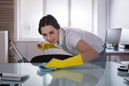 Happy Young Female Janitor Cleaning Desk With Rag At Workplaceの写真素材