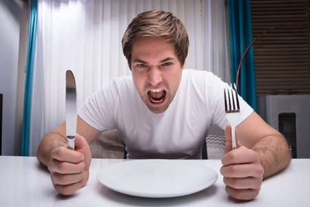 Angry Man Holding Knife And Fork With Empty Plate On Tableの写真素材