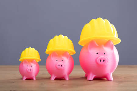 Close-up Of Increasing Pink Piggy Banks With Yellow Hard Hat On Wooden Deskの写真素材