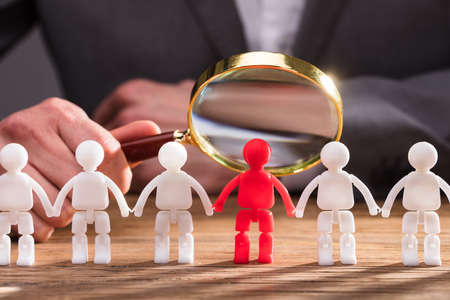 Close-up Of A Businessperson's Hand Holding Magnifying Glass Over Red Human Figure On Wooden Deskの写真素材