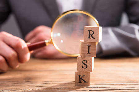 Close-up Of Wooden Blocks With Risk Word In Front Of Businessperson's Hand Holding Magnifying Glassの写真素材
