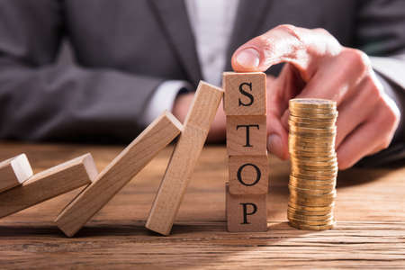 Human Finger Stopping Dominos From Falling With Stacked Coins On Wooden Deskの写真素材
