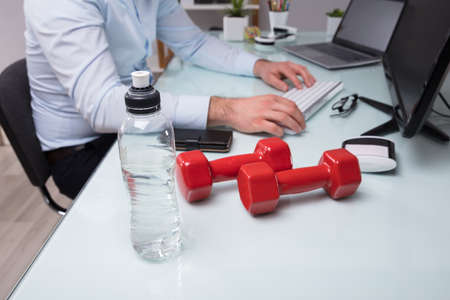 Close-up Of Water Bottle And Red Dumbbell In Front Of Businessperson Working In Officeの写真素材