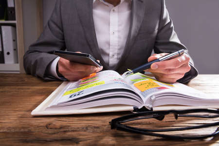 Businessperson's Hand Writing Schedule In Diary With Pen On Wooden Desk