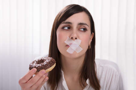 Close-up Of A Young Woman With Sticky Tape Over Her Mouth Unable To Eat Donutの写真素材