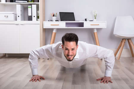 Portrait Of A Smiling Young Businessman Doing Push Up On Floor In Officeの写真素材
