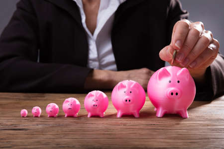 Close-up Of A Businessperson's Hand Inserting Coin In Piggybank Over Wooden Deskの写真素材